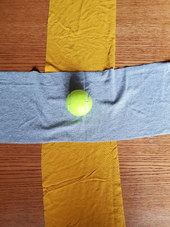 A tennis ball on top of two tshirt pieces that are in the shape of a cross.