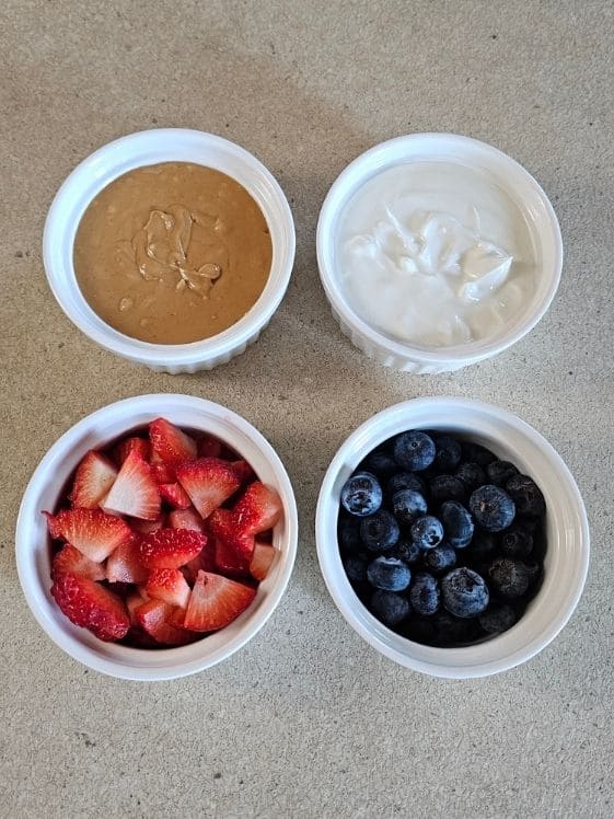 Peanut butter, Greek yogurt, strawberries, and blueberries in individual white bowls on the counter.