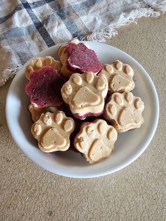 Paw shaped frozen peanut butter and jelly dog treats in a white bowl next to a linen towel.
