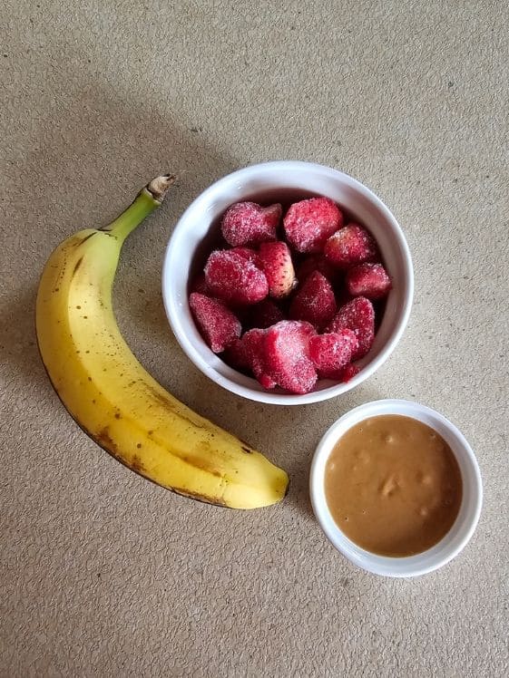 A banana next to a bowl of sliced strawberries and a bowl of peanut butter.