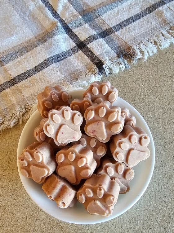 Paw shaped strawberry banana frozen dog treats in a white bowl next to a linen towel.