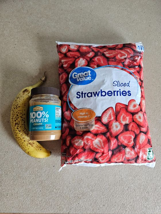 A ripe banana next to a jar of peanut butter that is next to a bag of frozen sliced strawberries.