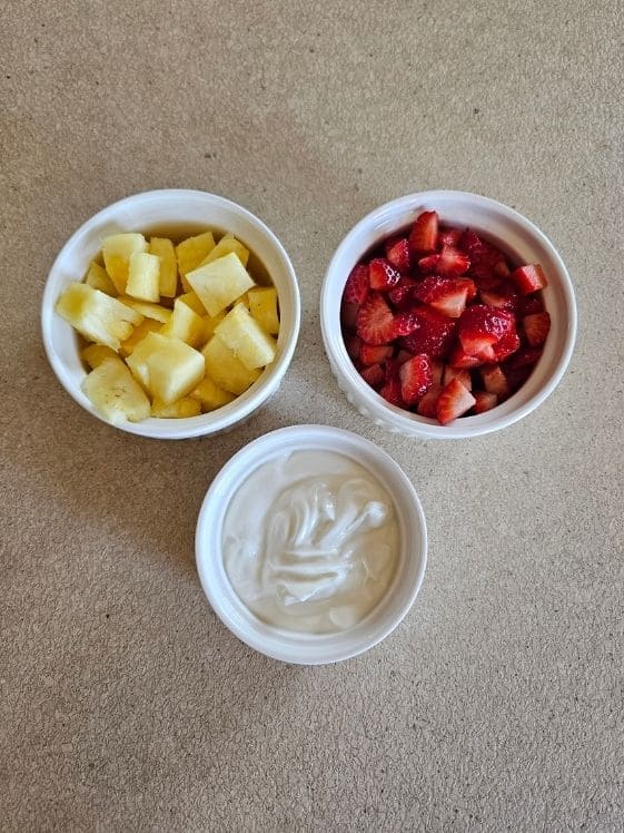 Pineapple chunks, diced strawberries, and Greek yogurt in individual white bowls.