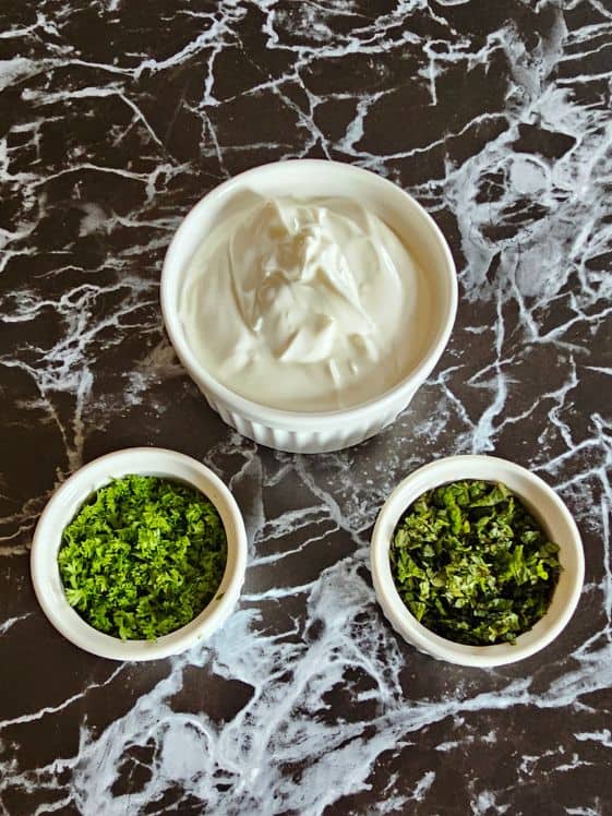 Greek yogurt, chopped mint leaves, and chopped parsley in individual white bowls on a dark marble surface.