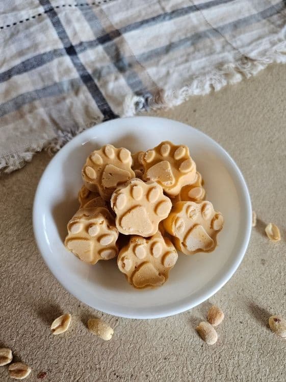 Paw shaped peanut butter sweet potato frozen dog treats in a white bowl next to a linen towel.