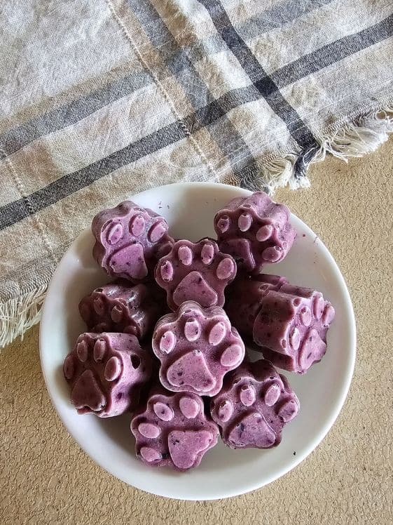 Paw shaped zucchini blueberry frozen dog treats in a white bowl next to a linen towel.