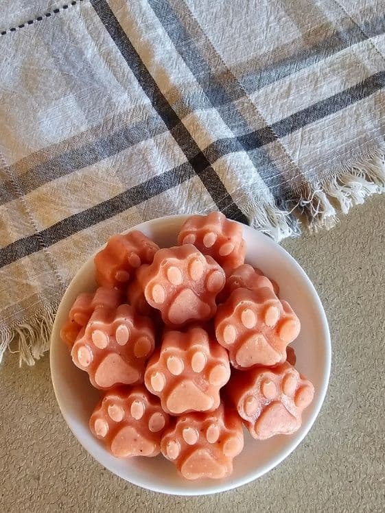 Paw shaped strawberry mango banana frozen dog treats in a white bowl next to a linen towel.