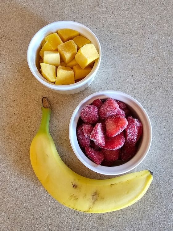 Cubed mango and sliced strawberries in individual white bowls next to a banana.