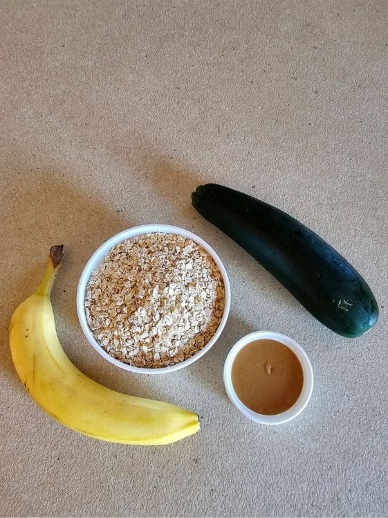 A banana, bowl of quick oats, a zucchini, and bowl of peanut butter on a counter top.