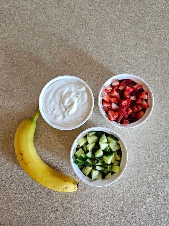 Greek yogurt, chopped strawberries, and chopped cucumbers in individual white bowls next to a banana.