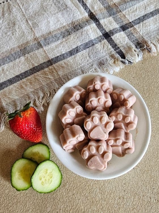Paw shaped berry cool cucumber pupsicles in a white bowl next to a strawberry, cucumber slices, and a linen towel.
