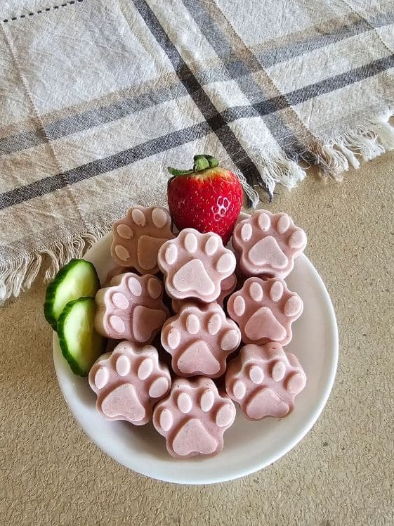 Paw shaped frozen strawberry cucumber dog treats in a white bowl with a strawberry and cucumber slices. A linen towel is next to the bowl.