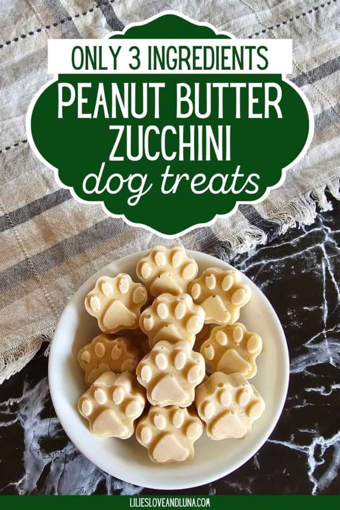 A bowl of paw-shaped frozen dog treats made from peanut butter and zucchini sits on a textured surface, labeled as "Only 3 Ingredients Peanut Butter Zucchini Dog Treats."