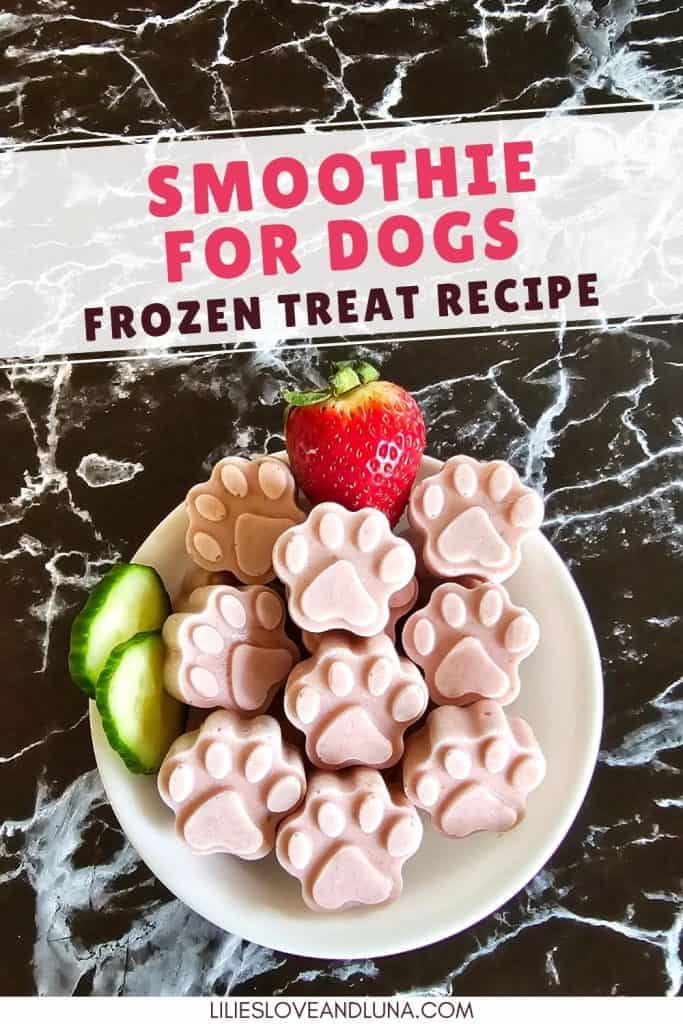 A plate of paw-shaped frozen dog treats made with strawberry yogurt is displayed alongside a fresh strawberry and cucumber slices on a marble surface.
