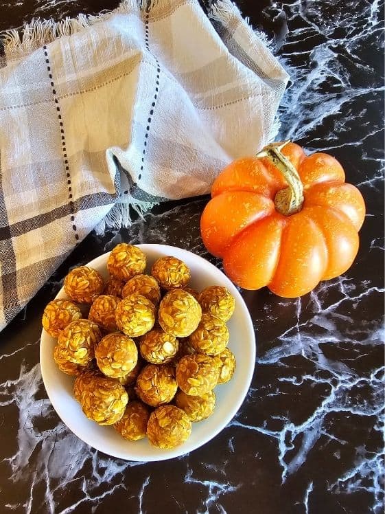 A bowl of no bake peanut butter pumpkin balls next to a small pumpkin and a linen towel.