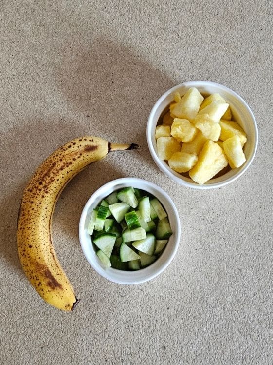 Frozen pineapple and chopped cucumbers in individual white bowls next to a ripe banana.